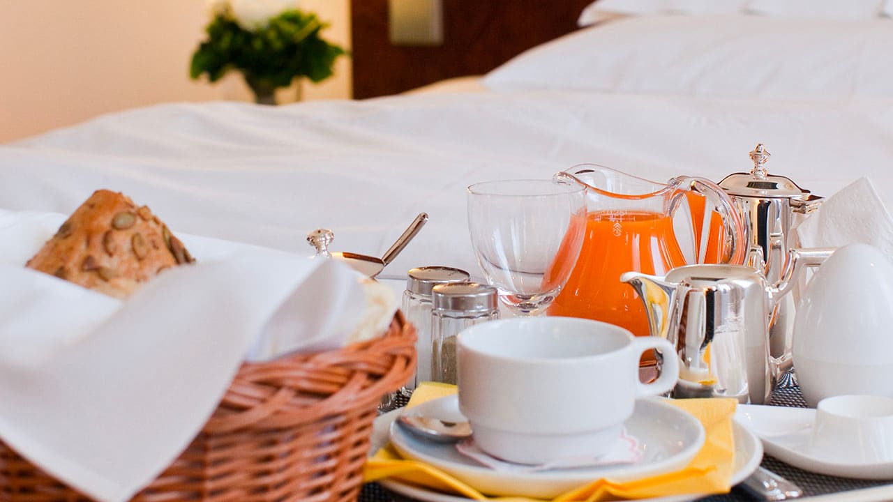 image-breakfast Frühstück auf einem Tablett im Hotelzimmer: Brötchen im Korb, Kaffee, Orangensaft, gekochtes Ei und Geschirr auf einem Bett serviert.