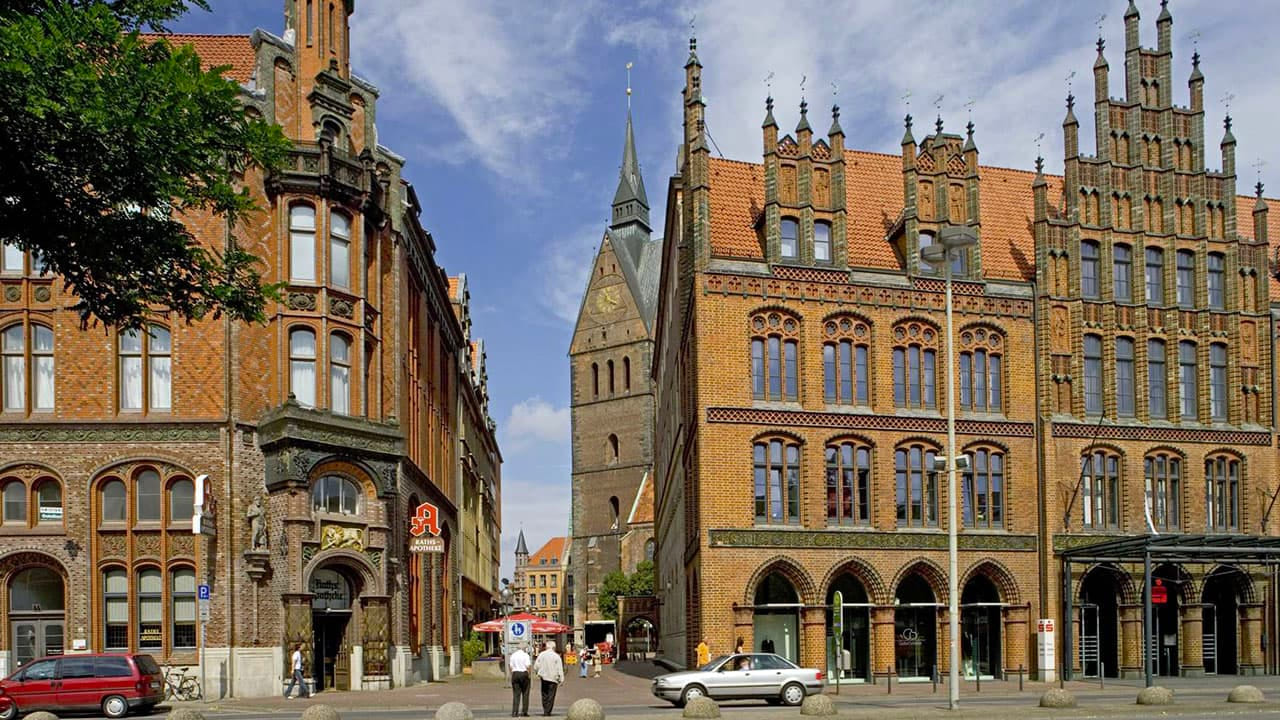 Historische Backsteinfassaden in der Altstadt von Hannover bei Tag. In der Mitte ragt der Turm der Marktkirche empor, flankiert von zwei reich verzierten Gebäuden im Stil der norddeutschen Backsteingotik.