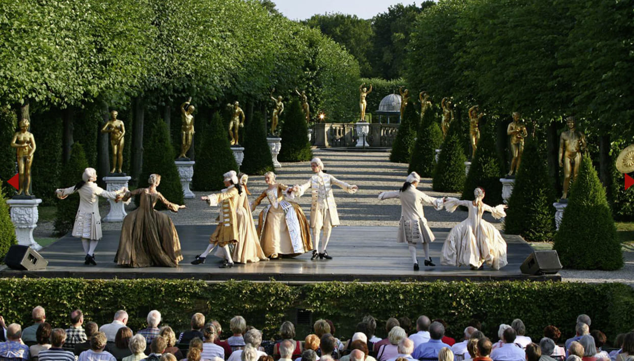 Barocktanz-Aufführung im Großen Garten Herrenhausen vor Publikum, mit goldenen Statuen und geschnittenen Bäumen im Hintergrund.