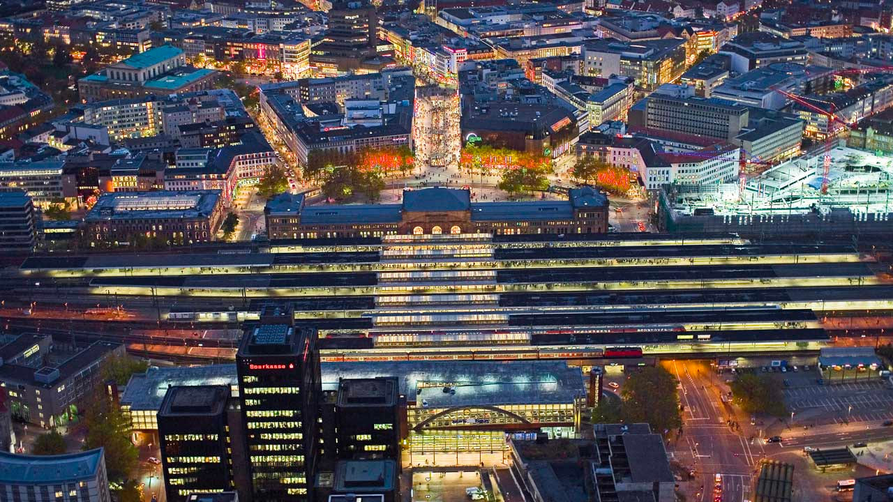 image-hannover-at-night Luftaufnahme des Hauptbahnhofs Hannover bei Nacht, hell beleuchtete Bahnsteige und umliegende Innenstadt mit Lichtern und Menschen.
