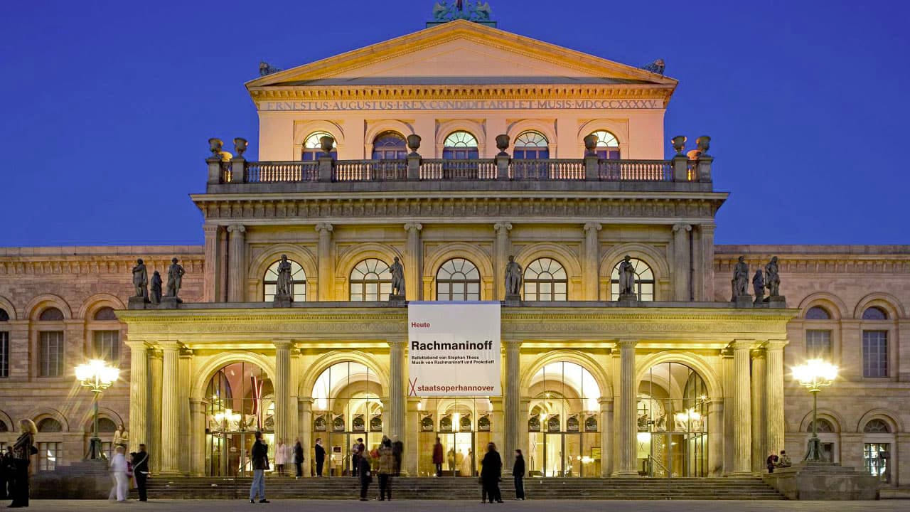 DE: Beleuchtete Staatsoper Hannover bei Abenddämmerung, Menschen stehen und gehen davor. / EN: Illuminated Hanover State Opera at dusk, with people standing and walking in front.