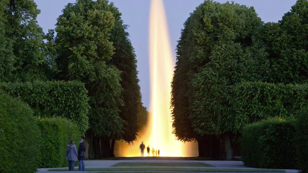 DE: Abendszene im Park mit hohen, dicht geschnittenen Bäumen. In der Mitte schießt eine große, beleuchtete Wasserfontäne in den Himmel, Menschen spazieren davor. / EN: Evening scene in a park with tall, neatly trimmed trees. In the center, a large illuminated water fountain shoots into the sky, with people walking in front of it.