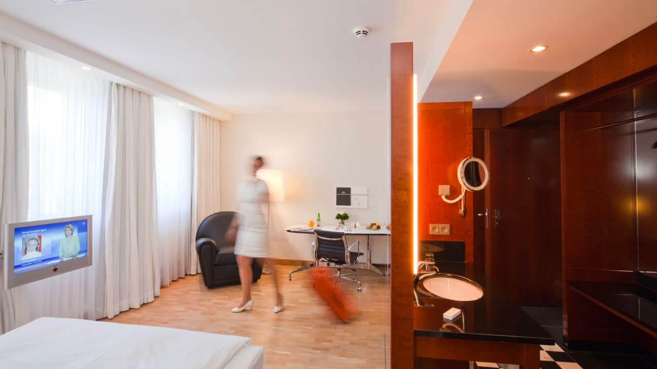 image-room-detail0 Modernes Einzelzimmer mit Bett, Fernseher, Schreibtisch und Sessel; im Vordergrund Waschtisch, im Hintergrund eine Frau mit Koffer in Bewegung.