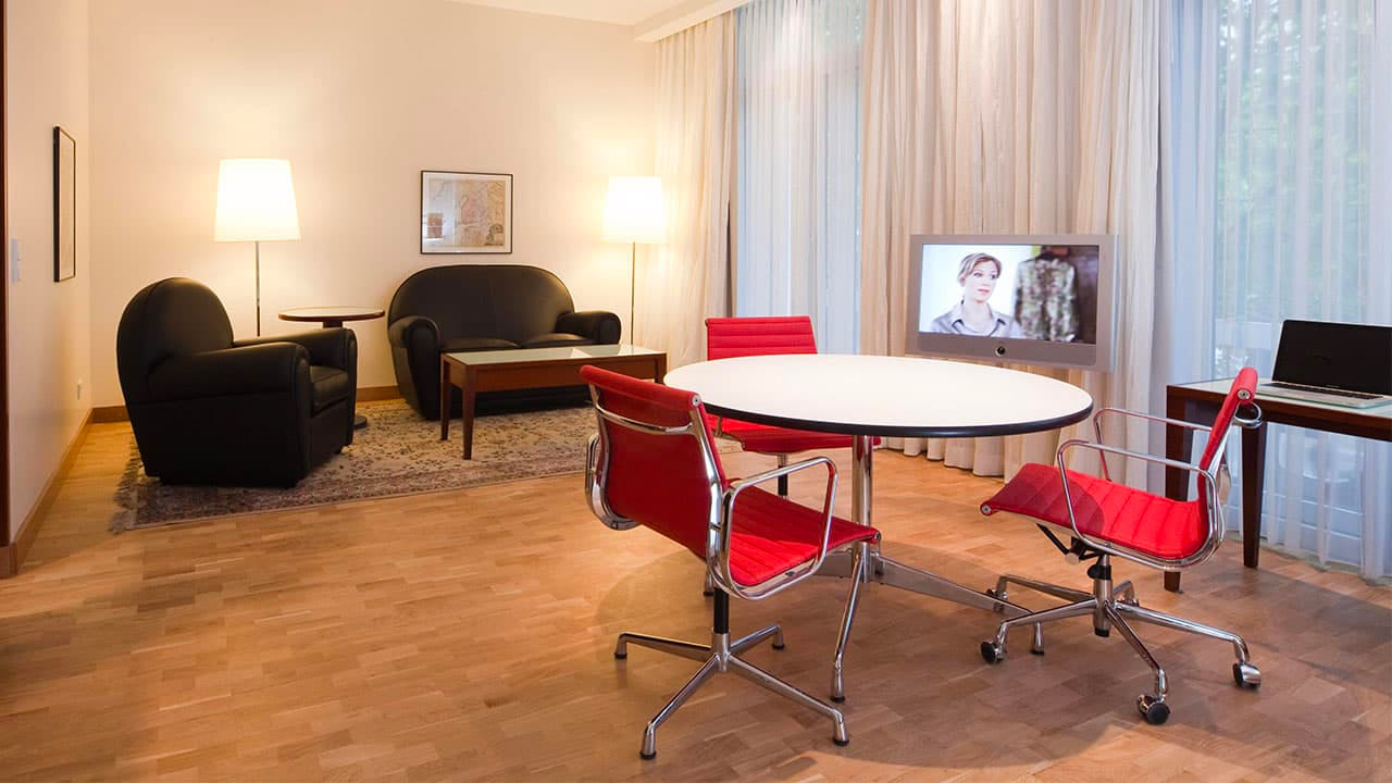 image-room-detail1 Wohnbereich einer Hotelsuite mit schwarzem Sofa, Sesseln, rundem Tisch mit roten Stühlen, Fernseher und Schreibtisch am Fenster.