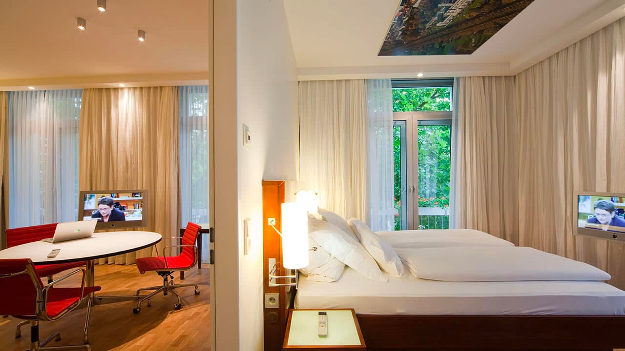 image-room-overview Blick in eine moderne Hotelsuite mit Schlafzimmer und separatem Arbeitsbereich, ausgestattet mit roten Designerstühlen, Schreibtisch und zwei Fernsehern.
