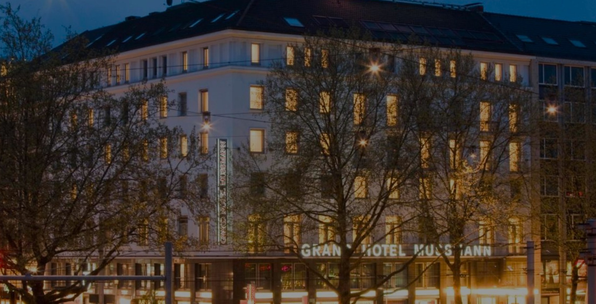 aufmacher-01 Außenansicht des Grand Hotel Mussmann am Abend, beleuchtete Fenster und Hotel-Schriftzug, Bäume im Vordergrund, dunkler Himmel mit Straßenbeleuchtung.