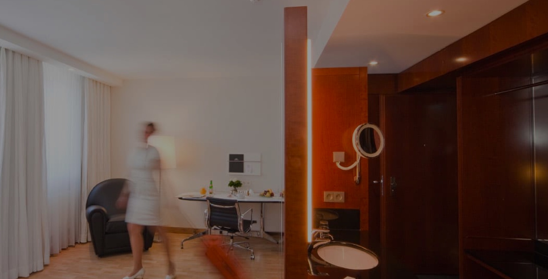 aufmacher-02 Modernes Hotelzimmer mit hellen Vorhängen, Schreibtisch und Sessel; im Vordergrund unscharfe Person in Bewegung, Spiegel und Waschbereich mit dunkler Holzverkleidung sichtbar.
