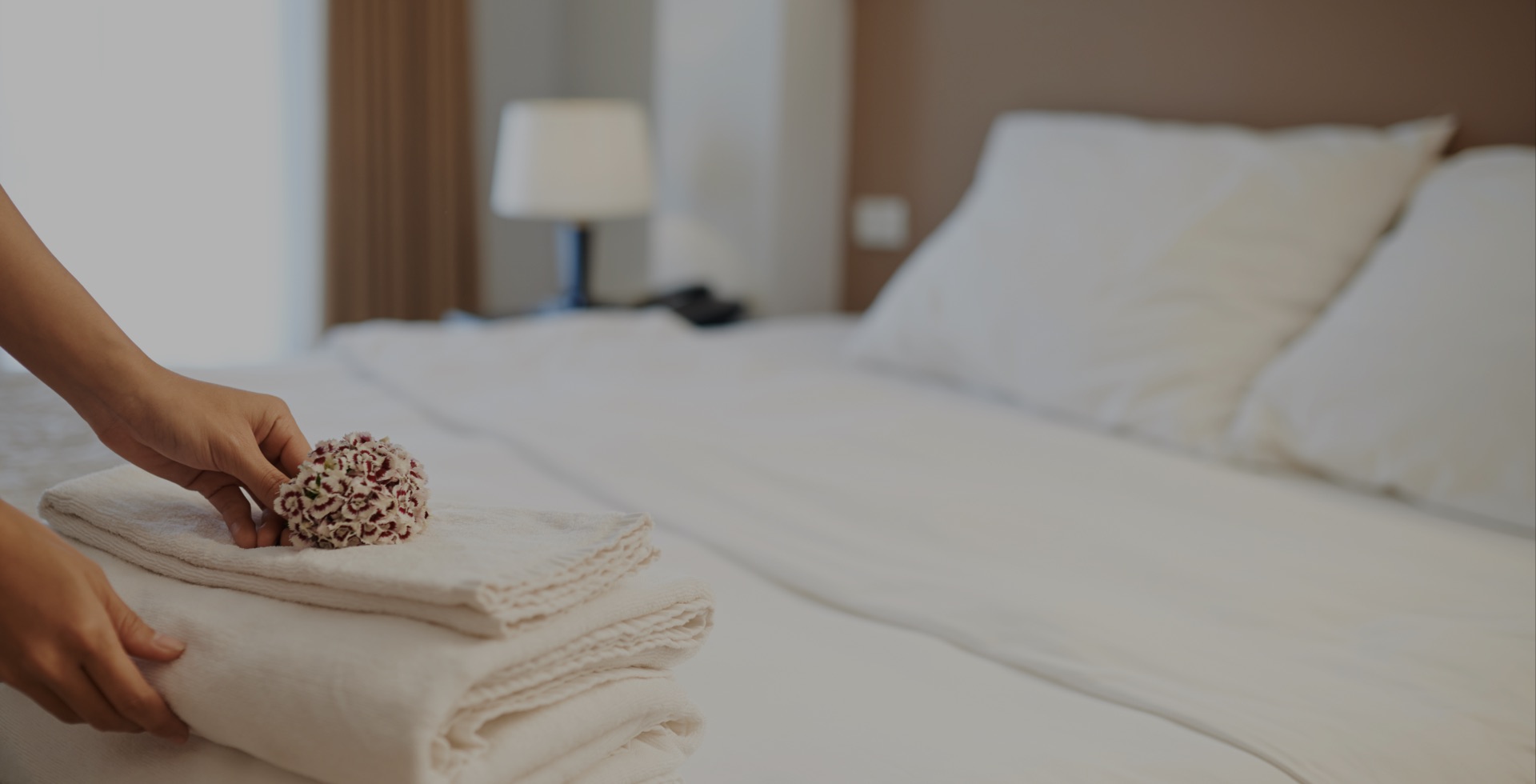 aufmacher-03 Nahaufnahme eines gemachten Hotelbetts, auf dem eine Hand frische Handtücher mit einer dekorativen Blume ablegt, im Hintergrund eine Nachttischlampe.