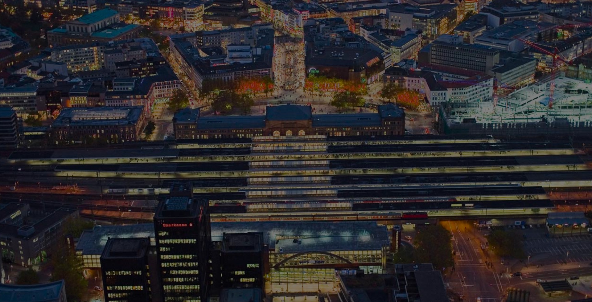 aufmacher-04 Abendliche Luftaufnahme des Hauptbahnhofs Hannover mit beleuchteten Bahnsteigen und belebter Innenstadt im Hintergrund.