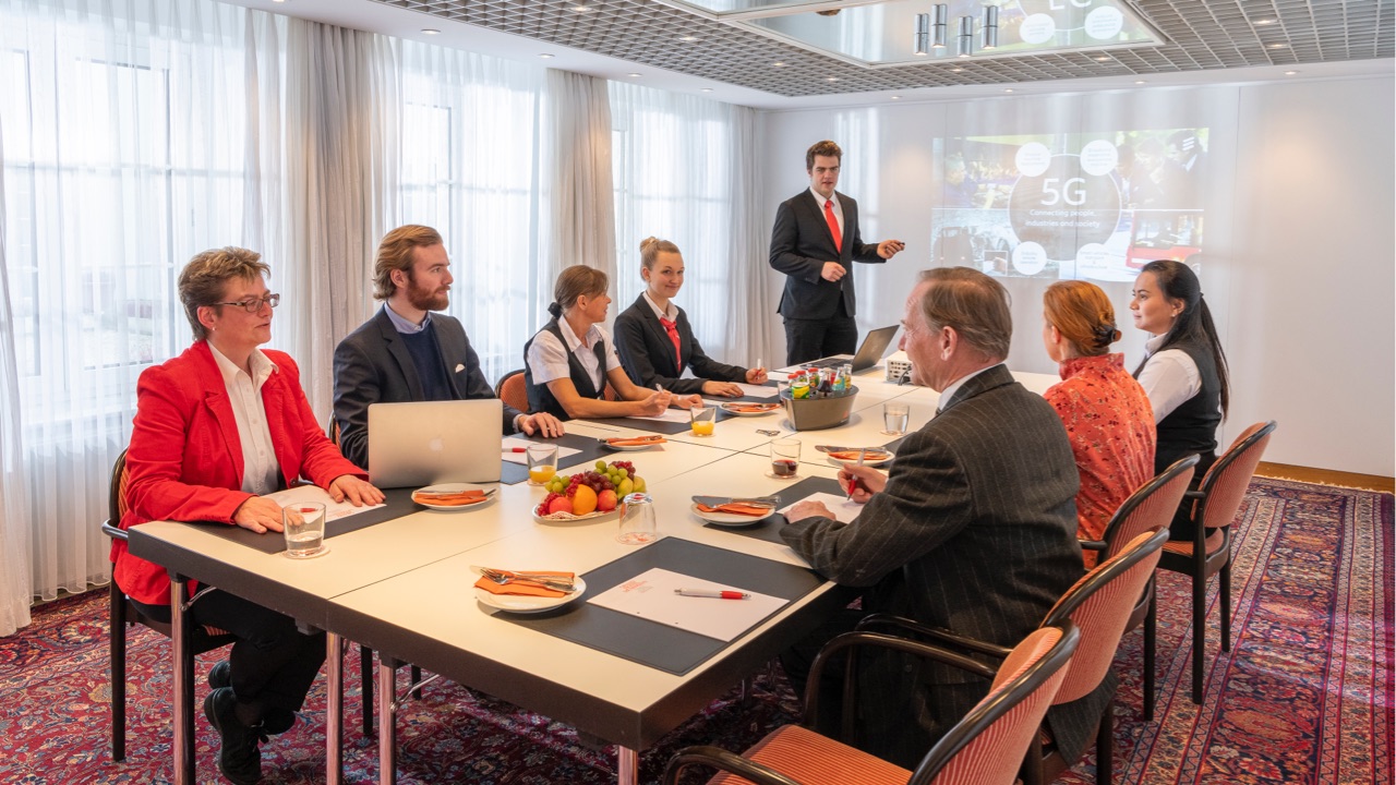 DE: Meeting mit acht Personen an einem Konferenztisch, Vortragender steht vorne vor einer Projektion zum Thema 5G, Teilnehmende in Businesskleidung, Tisch mit Getränken, Obst und Schreibmaterialien gedeckt. / EN: Meeting with eight people at a conference table, presenter standing at the front in front of a projection on the topic of 5G, participants in business attire, table set with drinks, fruit, and writing materials.
