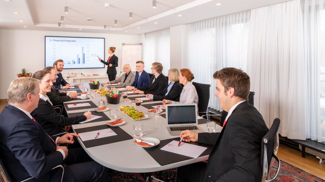 DE: Konferenzraum mit 11 Teilnehmenden in Businesskleidung, Vortragende präsentiert vor Bildschirm mit Finanzgrafik, rechteckiger Tisch mit Snacks, Getränken und Konferenzunterlagen, Tageslicht durch große Fenster. / EN: Conference room with 11 participants in business attire, presenter in front of a screen showing a financial graphic, rectangular table with snacks, drinks, and conference materials, daylight through large windows.