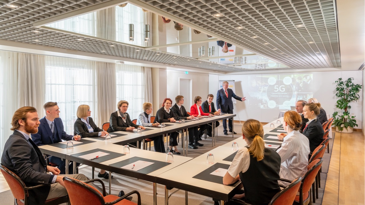 DE: U-förmig angeordneter Konferenztisch mit 15 Personen in Businesskleidung, Vortragender präsentiert vor einer Projektionswand mit 5G-Diagramm, heller Raum mit großer Fensterfront und moderner Decke mit Spiegelplatten. / EN: U-shaped conference table with 15 people in business attire, presenter in front of a projection screen with a 5G diagram, bright room with large windows and a modern ceiling with mirrored panels.