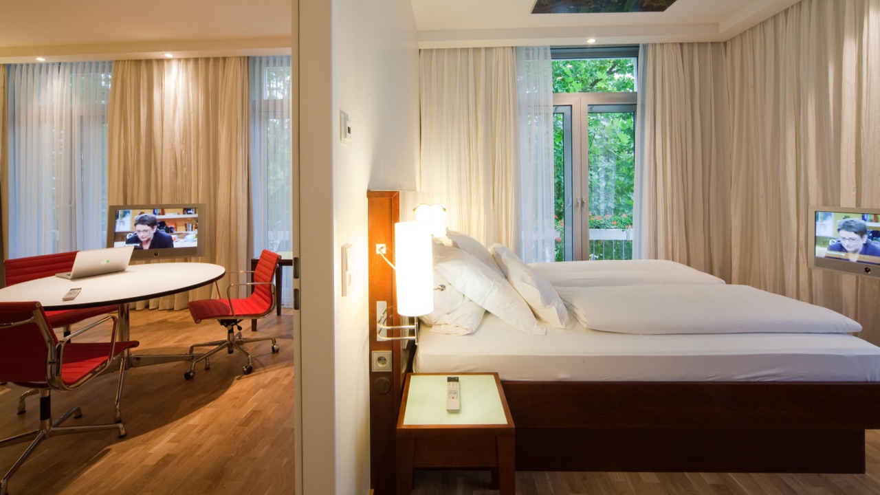 DE: Blick in eine moderne Hotelsuite mit Schlafzimmer und separatem Arbeitsbereich, ausgestattet mit roten Designerstühlen, Schreibtisch und zwei Fernsehern. / EN: View into a modern hotel suite with a bedroom and separate work area, featuring red designer chairs, a desk, and two TVs.