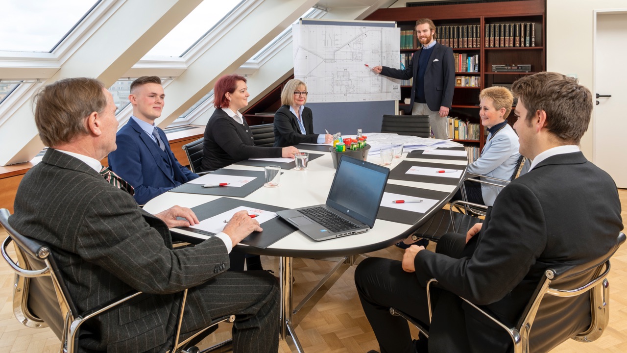Geschäftliches Meeting mit sieben Personen an einem Konferenztisch, ein Mann präsentiert Baupläne auf einem Flipchart.