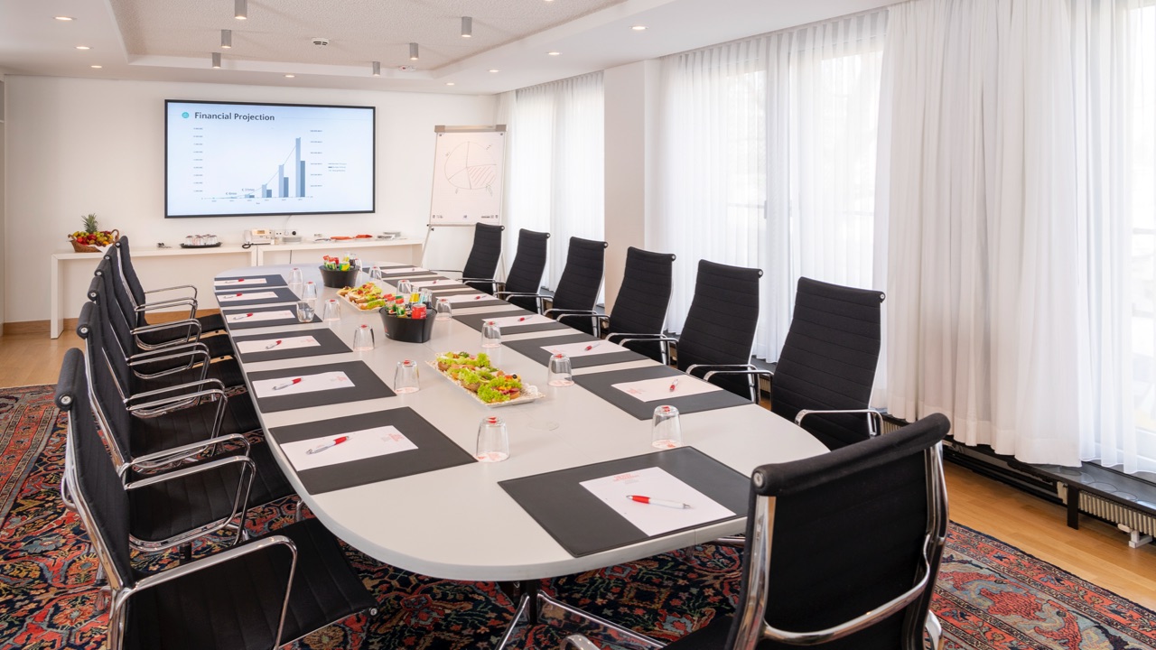 Moderner Konferenzraum mit großem Besprechungstisch und 13 schwarzen Lederstühlen, Präsentation mit Finanzdiagramm auf Bildschirm, Flipchart, Tageslicht durch bodentiefe Fenster, Snacks und Getränke auf dem Tisch.
