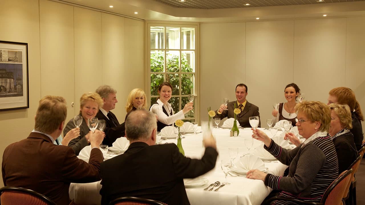Grand-Hotel-Mussmann-Veranstaltung-Georg-01 Gesellige Runde von 13 Personen beim Anstoßen an einem festlich gedeckten Tisch in einem Veranstaltungsraum.