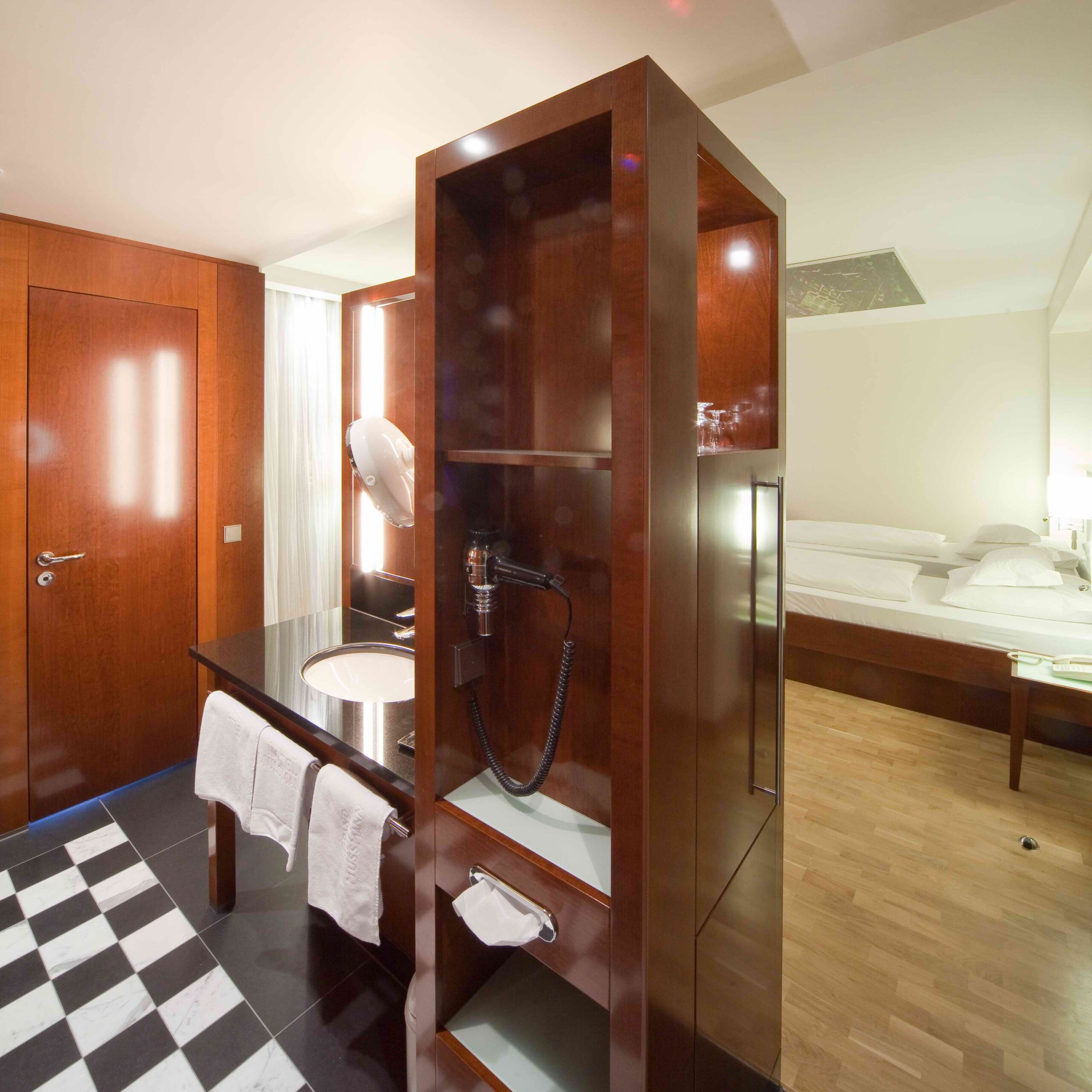 DE: Hotelzimmer mit Doppelbett und angrenzendem Badbereich mit beleuchtetem Spiegel, Waschbecken und Marmorboden. / EN: Hotel room with a double bed and adjoining bathroom area with illuminated mirror, sink, and marble floor.
